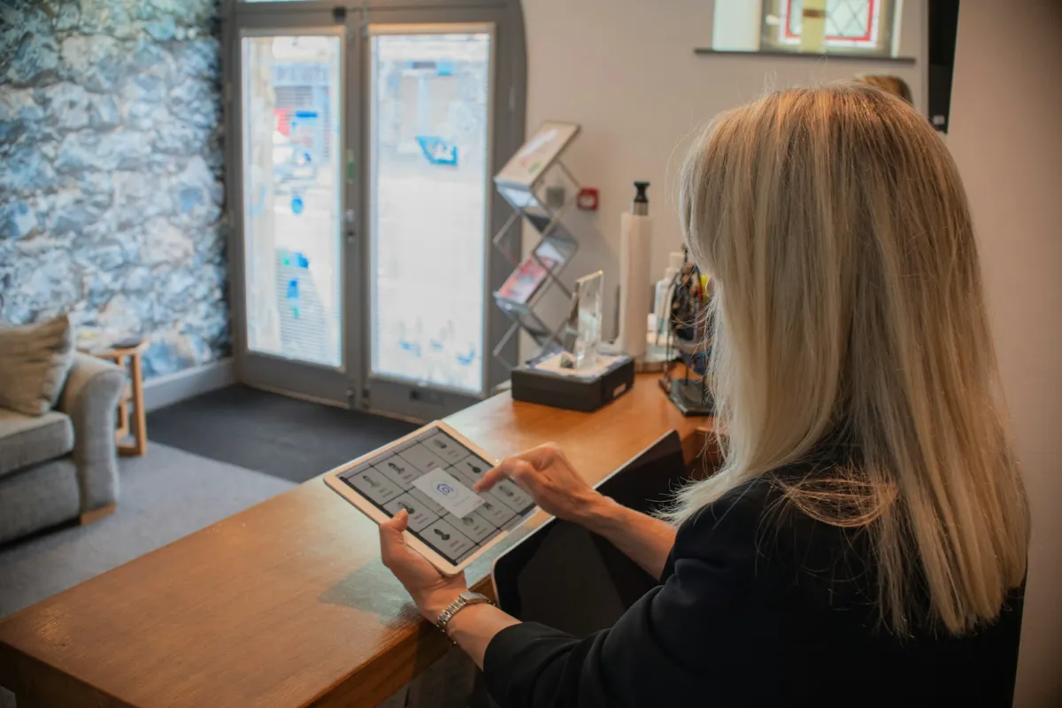 Receptionist at a dental practice reception desk using a tablet, with the waiting area behind her